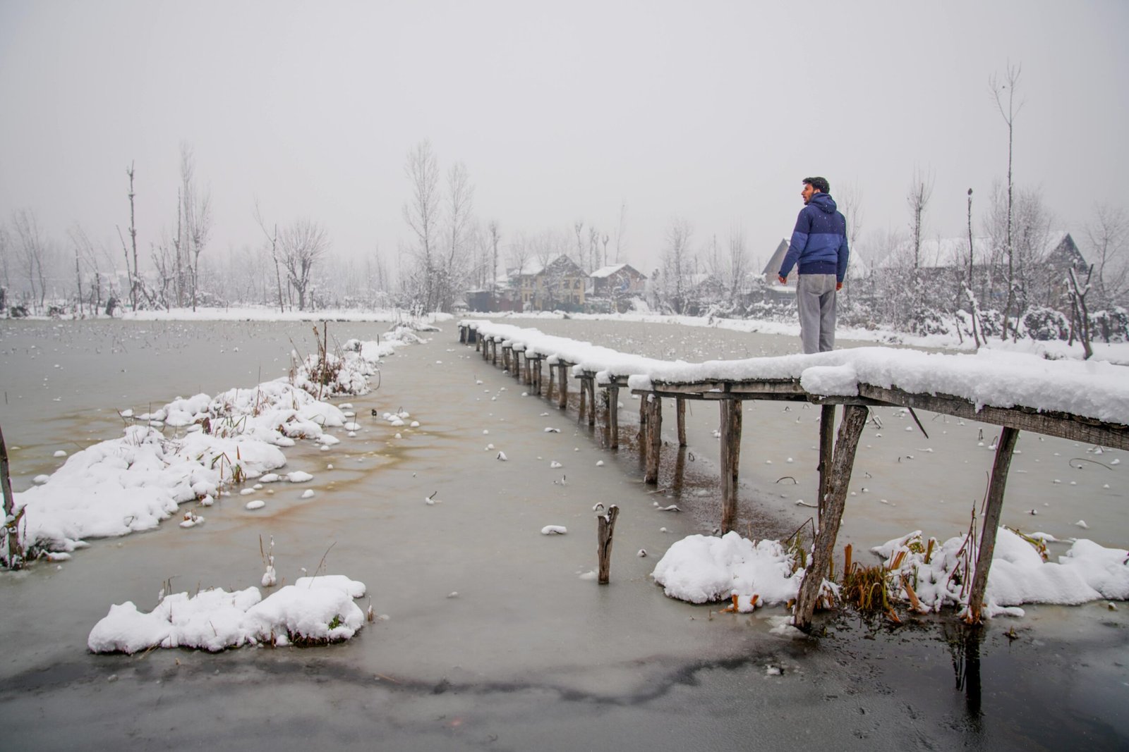 A serene winter scene in Kashmir with a snow-covered bridge and a person gazing into the distance.
