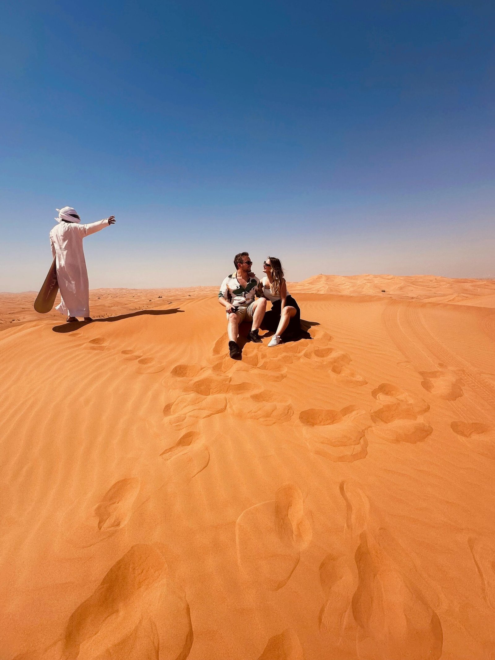 Couple sitting on sand dunes in Dubai desert with guide in traditional attire.